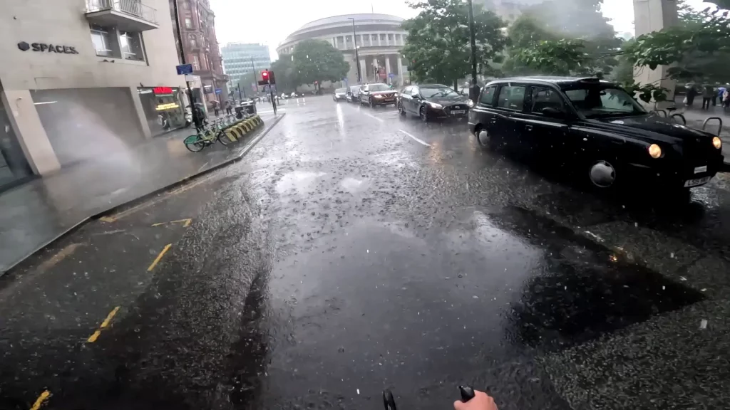 A view from a cyclist of a very rainy day on a busy town road, the water has coated the road and you can see the rain drops hitting of the wet ground.