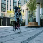 Image shows a delivery cyclist with a take out food backpack riding through and empty plaza.
